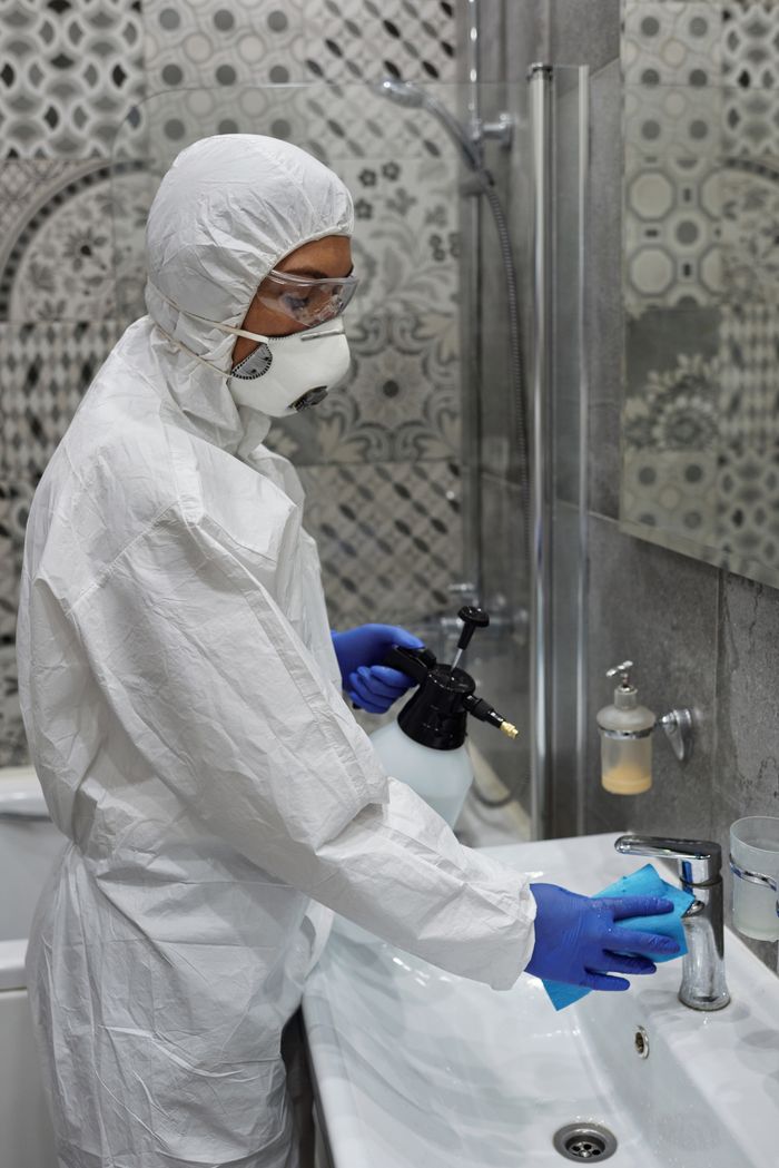 Person in full PPE cleans a bathroom sink with spray bottle and cloth for hygiene.